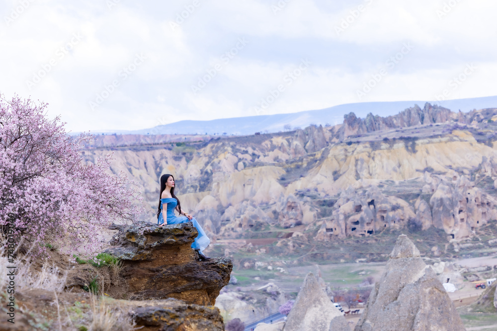 Beautiful woman in blue dress on Turkey rocks mountain.Cheerful woman relax outdoor with beautiful sky and pink flowers in holiday vacation.Woman tourist enjoy amazing mountain view.Travel and freedom