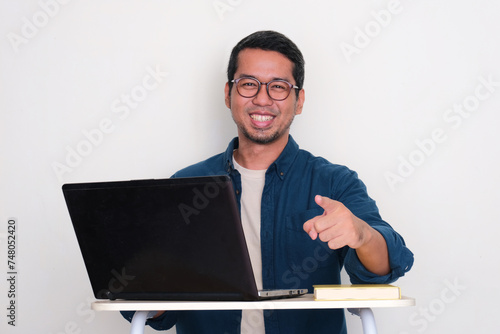 Adult Asian man sitting in front of a laptop smiling and pointing at the camera photo