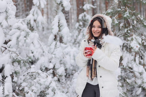 Młoda ładna dziewczyna trzyma w dłoniach czerwony kubek z gorącą herbatą. Spaceruje w mroźny zimowy dzień po lesie.