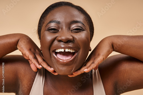 cheerful plus size african american woman smiling with hands near her face on beige background