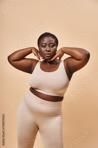 Plus size African American woman in beige underwear posing with hands near face on beige background