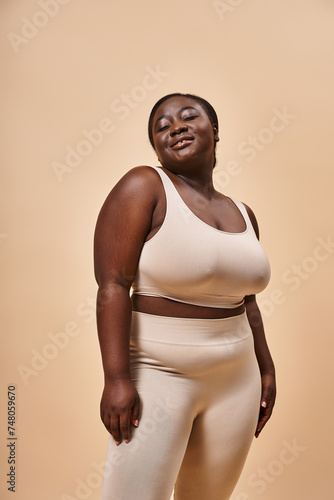 Radiant plus size african american woman in beige sportswear smiling on matching backdrop