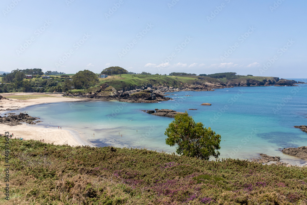 a serene beach scene with calm, turquoise waters, a sandy shore bordered by rocky outcrops and lush greenery under a clear sky. A few people are visible at a distance