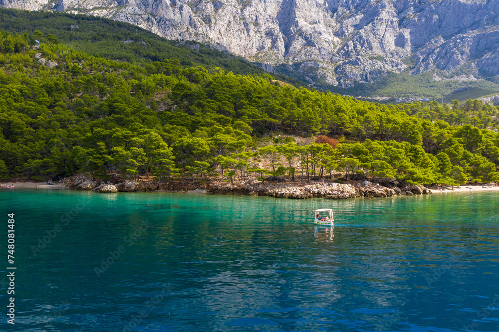 A small boat with tourists slowly floats on clear water. Amazing coastline. Aerial photography. The mountains are covered with greenery, the blue transparent sea. Makarska Riviera. Croatia