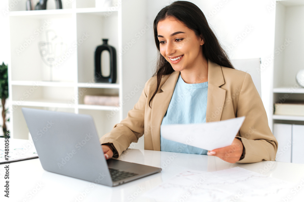 Successful indian or arabian business woman, ceo, financial manager, secretary, sit at workplace in modern office with laptop, studying documents, analyzing business plan and strategy, smiling Successful indian or arabian business woman, ceo, financial manager, secretary, sit at workplace in modern office with laptop, studying documents, analyzing business plan and strategy, smiling