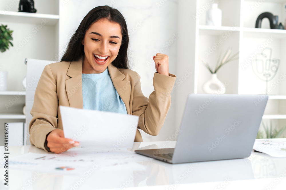Joyful indian or arabian business woman, ceo, financial manager, secretary, sit at workplace in modern office with laptop, studying documents, rejoicing in a good results, gesturing hand, smiling Joyful indian or arabian business woman, ceo, financial manager, secretary, sit at workplace in modern office with laptop, studying documents, rejoicing in a good results, gesturing hand, smiling