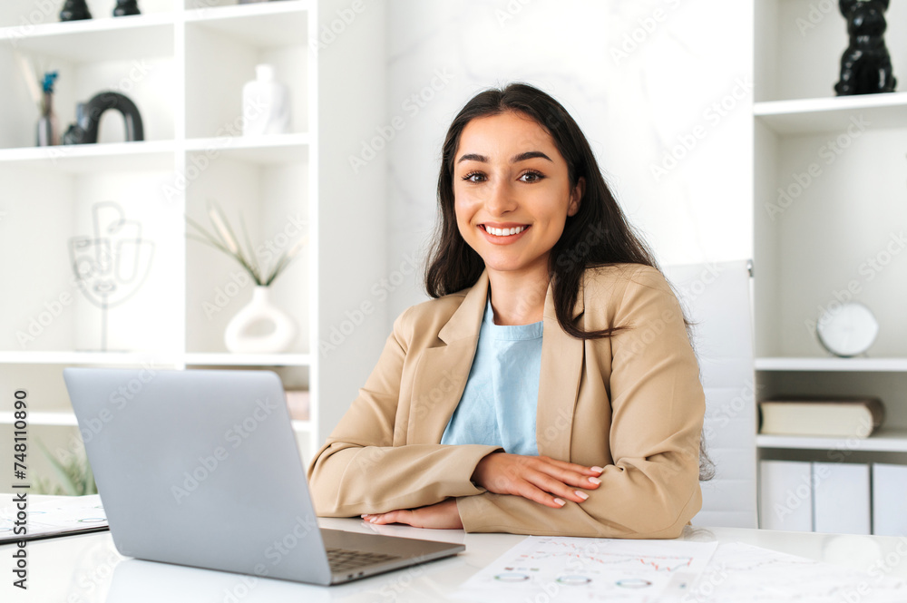 Beautiful positive successful indian or arabian business woman, company digital specialist, sitting at workplace in the office with laptop, working on a project, looks at camera, smiling friendly Beautiful positive successful indian or arabian business woman, company digital specialist, sitting at workplace in the office with laptop, working on a project, looks at camera, smiling friendly