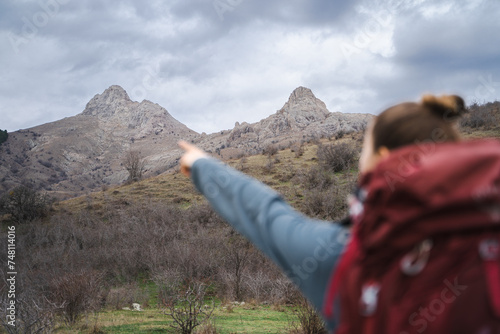 Back view of young traveler woman with red backpack points with finger at the mountains background, focus on the mountains, copy space
