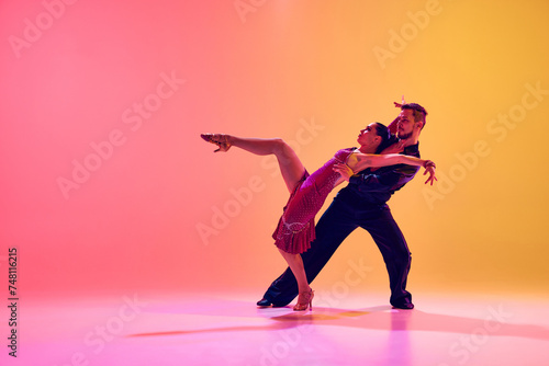 Fototapeta Naklejka Na Ścianę i Meble -  Young couple, man and woman in motion, ballroom dancers making creative performance against gradient pink yellow background in neon light. Concept of dance class, hobby, art, dance school, talent