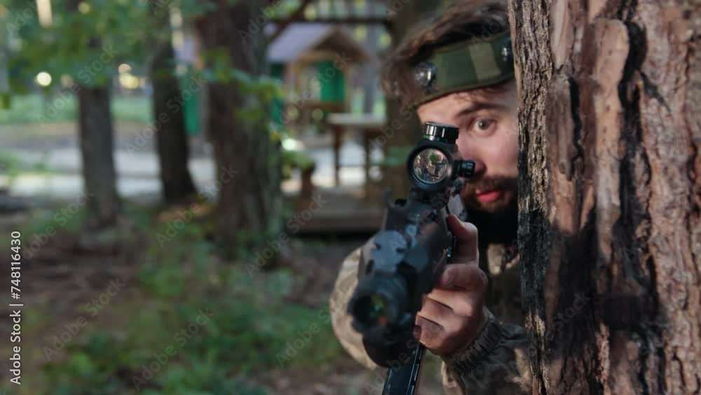 Aiming precision, tactical position, combat attire. In forest environment, man in camo clothing with marker on his head is positioned against tree, accurately targeting with Laser tag gun Aiming precision, tactical position, combat attire. In forest environment, man in camo clothing with marker on his head is positioned against tree, accurately targeting with Laser tag gun