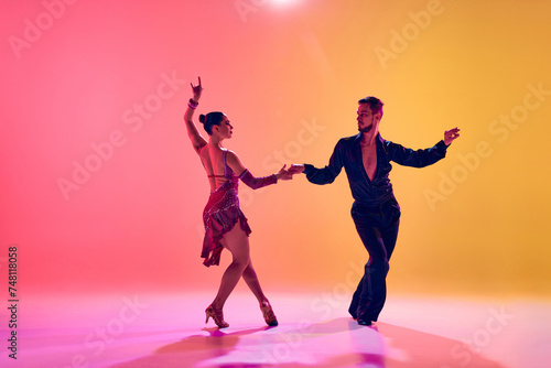 Male and female dancers performing a Latin dance in stylish clothes against gradient pink yellow background in neon light. Concept of dance class, hobby, art, dance school, talent