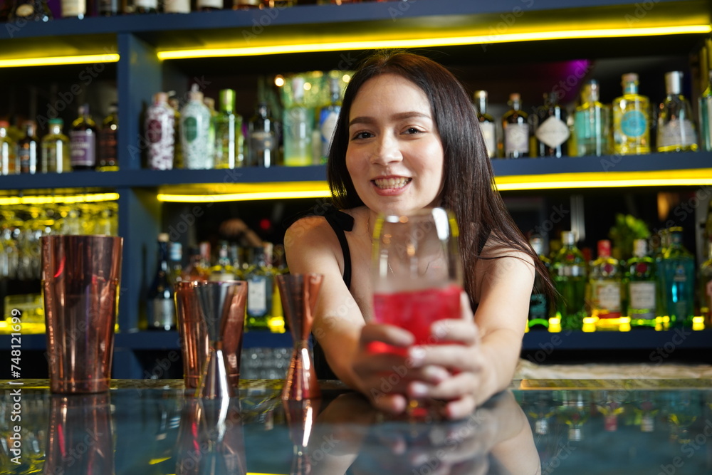 young Asian woman works as a bartender standing behind the counter holding a stainless steel shaker and mixing drinks for customers.