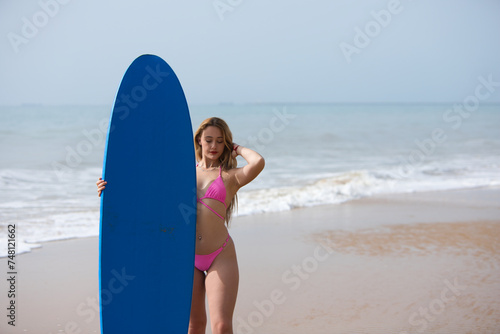 Young and beautiful blonde surfer woman in pink bikini and blue surfboard. The girl enjoys her holidays on the beach to practice her favourite sport. Travel and holiday concept