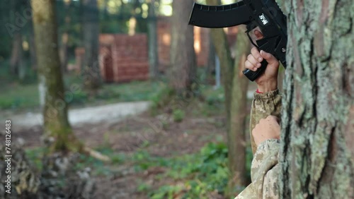 Tag shooter, precision aiming, enemy engagement. Man adorned in camouflage clothing with marker on his head leans against tree, skillfully targeting with Laser tag gun and firing at opponent photo