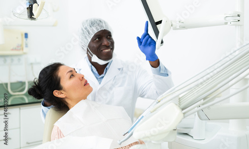 Positive african american dentist talking to asian woman during dental checkup, discussing teeth x-ray and treatment procedures