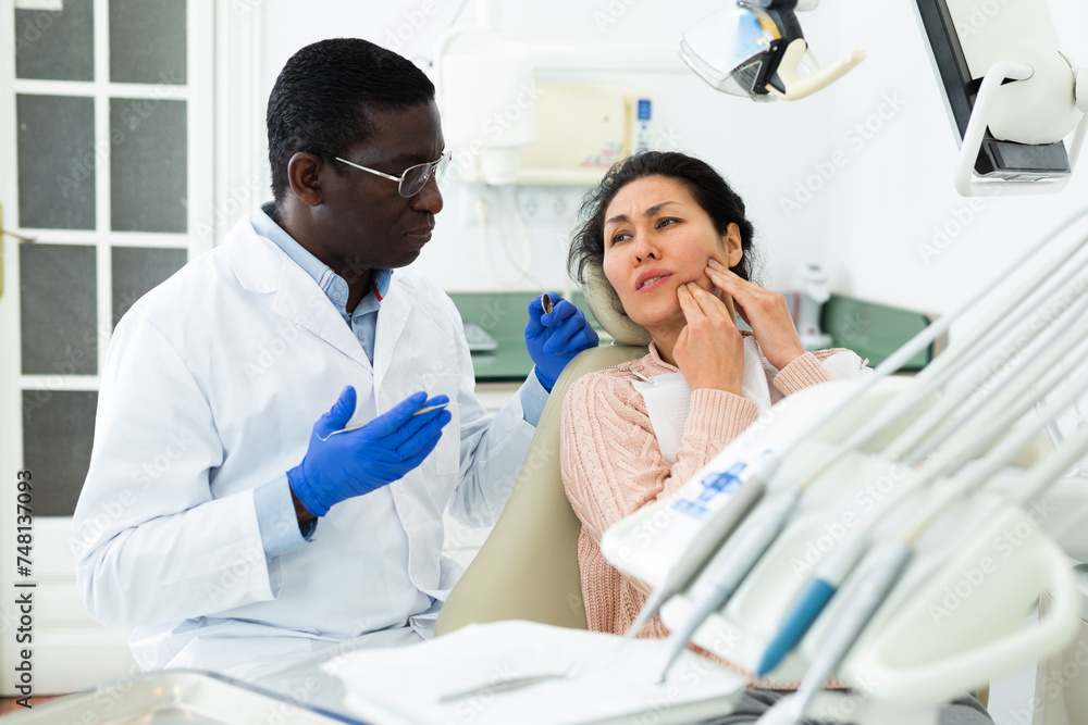 Fototapeta premium Asian woman complaining about toothache to african-american man dentist during consulatation in clinic.