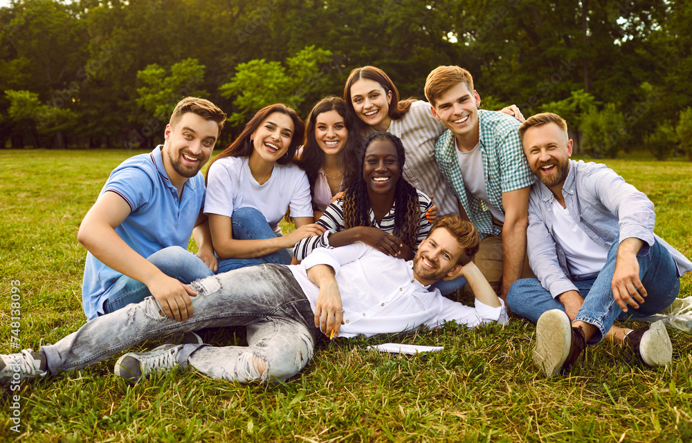 Fototapeta premium Portrait of a group of happy people friends sitting in the summer park hugging and smiling. Young students looking cheerful at camera outdoors. Friendship and togetherness concept. Banner.