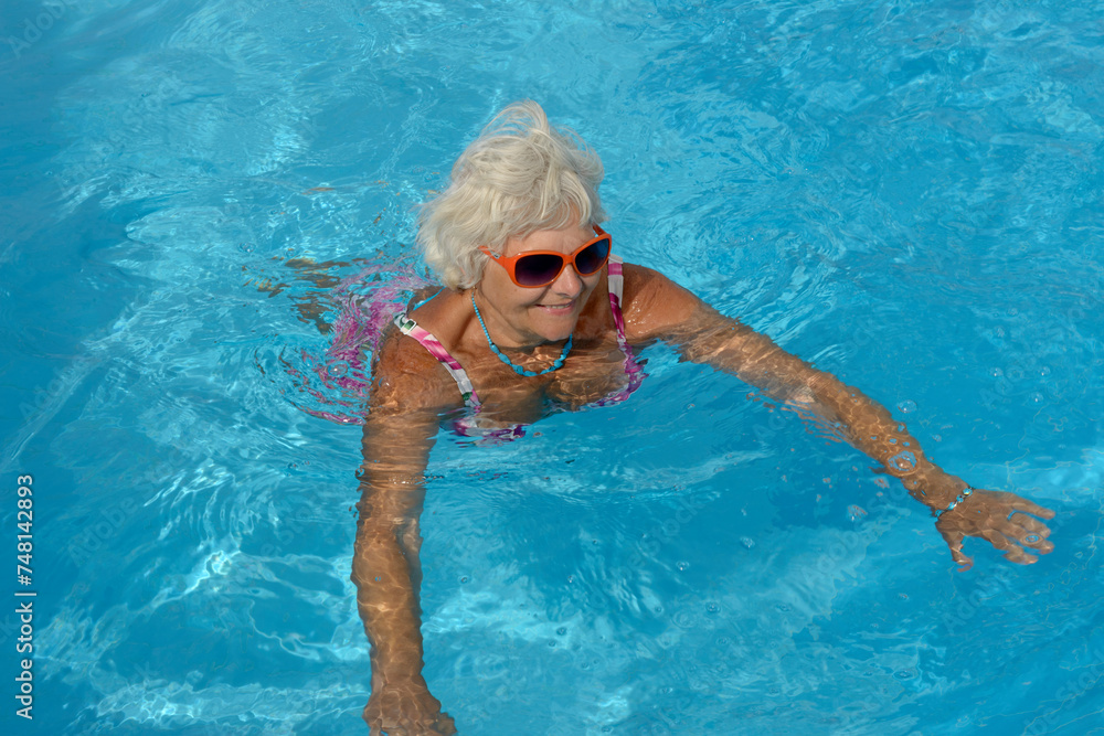 Aged woman is swimming in bright blue water of pool.