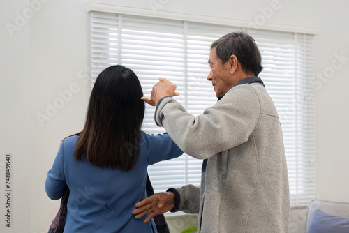 Elderly Asian couple in their 60s dancing in the living room together, enjoying music and smiling in love.