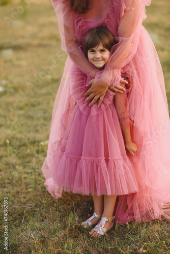 Mother and her daughter in the same designed pink dresses posing on nature at summer. Mum hugging her little girl. Love