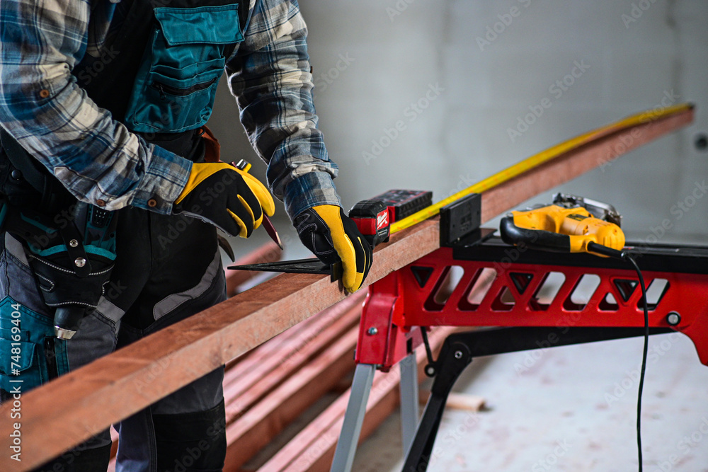 Fototapeta premium a worker in fitters and gloves with a tool belt measures wood using a tape measure and a right-angled triangle, blurred background