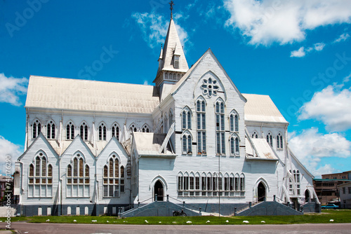 Schilderij op canvas A tall white wooden cathedral with a spire and elongated windows on a sunny day against a blue sky with clouds, Guyana