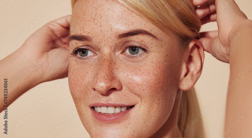 Naklejka premium Close up highly detailed portrait of a young smiling woman with freckles. Cropped image of a beautiful blond female adjusting hair against a beige backdrop.