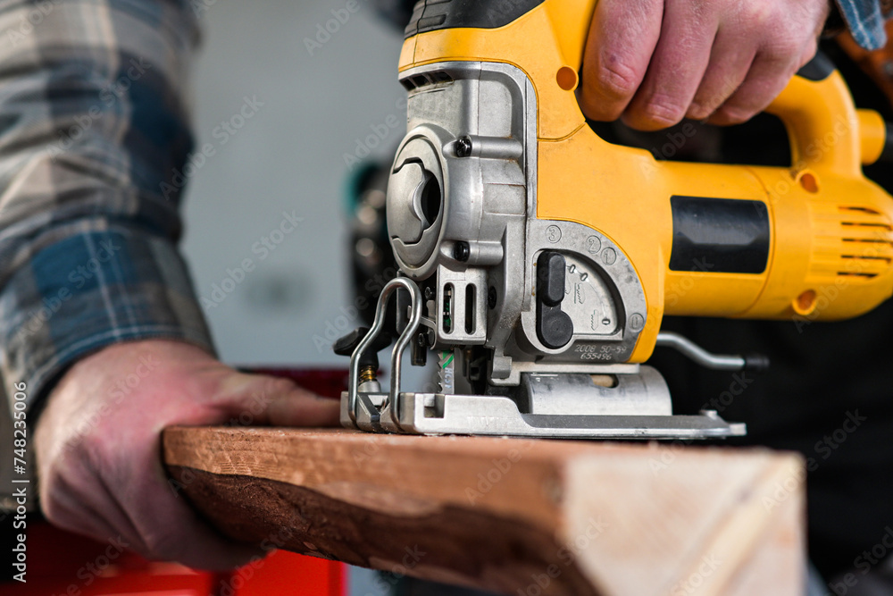 worker in overalls and wearing yellow gloves sawing wood with a reciprocating electric wood saw - close-up view and blurred background