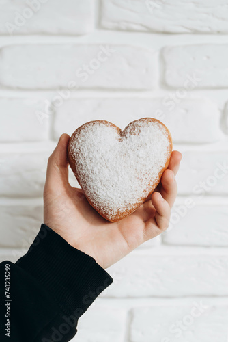 A heart-shaped donut sprinkled with powder in a man's hand close-up against a white brick wall. photo