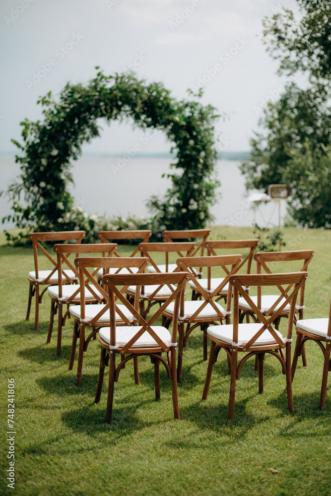 Wedding aisle with wooden chairs, natural, rustic wedding decor on a green meadow. Wedding ceremony with arch and wooden chairs, water view