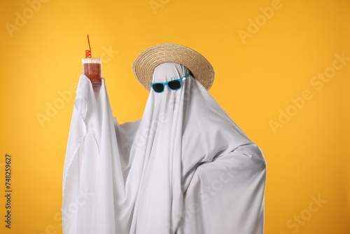 Person in ghost costume, sunglasses and straw hat holding glass of drink on yellow background