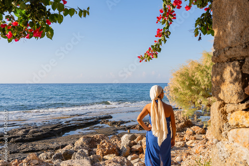 Beautiful woman in Greek dress and arms akimbo enjoying a sunrise by the sea and observing a bougainvillea in bloom in summer photo