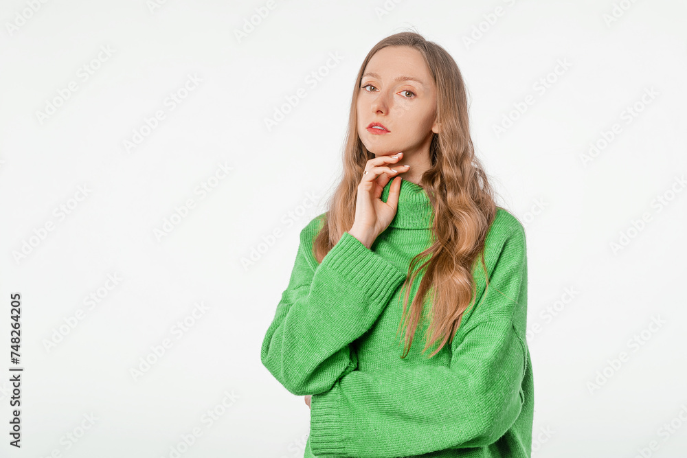 Portrait of young woman thinking, touching chin and looking skeptical at camera, standing in knitwear green sweater over white background Portrait of young woman thinking, touching chin and looking skeptical at camera, standing in knitwear green sweater over white background