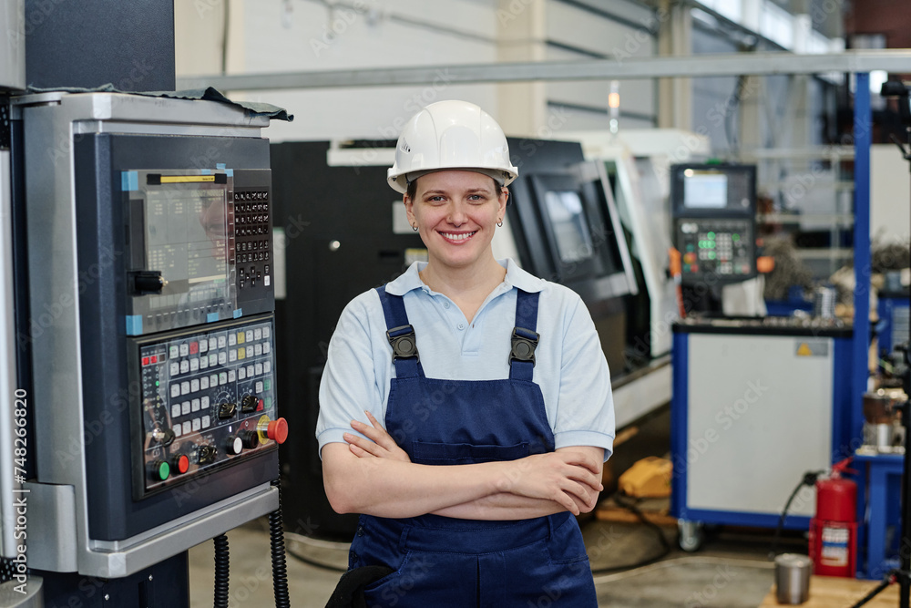 Medium portrait of self-confident female turner standing with arms crossed in factory workshop smiling at camera Medium portrait of self-confident female turner standing with arms crossed in factory workshop smiling at camera