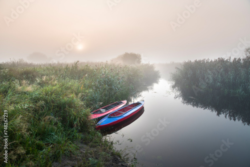Two multi-colored kayaks at sunrise near the river bank in the morning mist among the river grass.