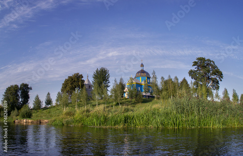 An old wooden beautiful Orthodox church among the trees on the banks of the river against a blue cloudy sky.