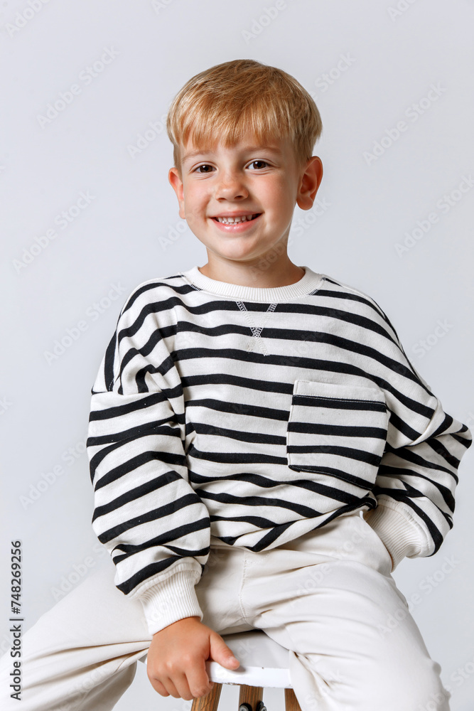 Blonde little kid in striped sweater and white trousers sitting posing at studio Blonde little kid in striped sweater and white trousers sitting posing at studio