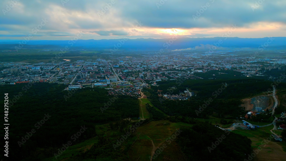 Aerial of the city and big green hills. Clip. Summer big city and cloudy sky during sunset.