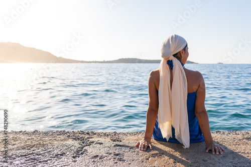 Beautiful close up of woman with bare back and headscarf by the sea sitting on a jetty at dawn in southern Crete