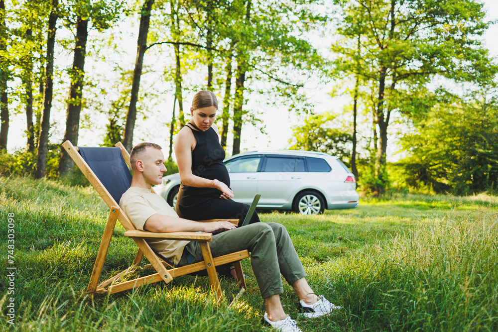 Fototapeta premium A family couple, a man and a woman, are relaxing in nature and working remotely with a laptop. Picnic of loving couples with work online. A pregnant woman rests in nature with her husband