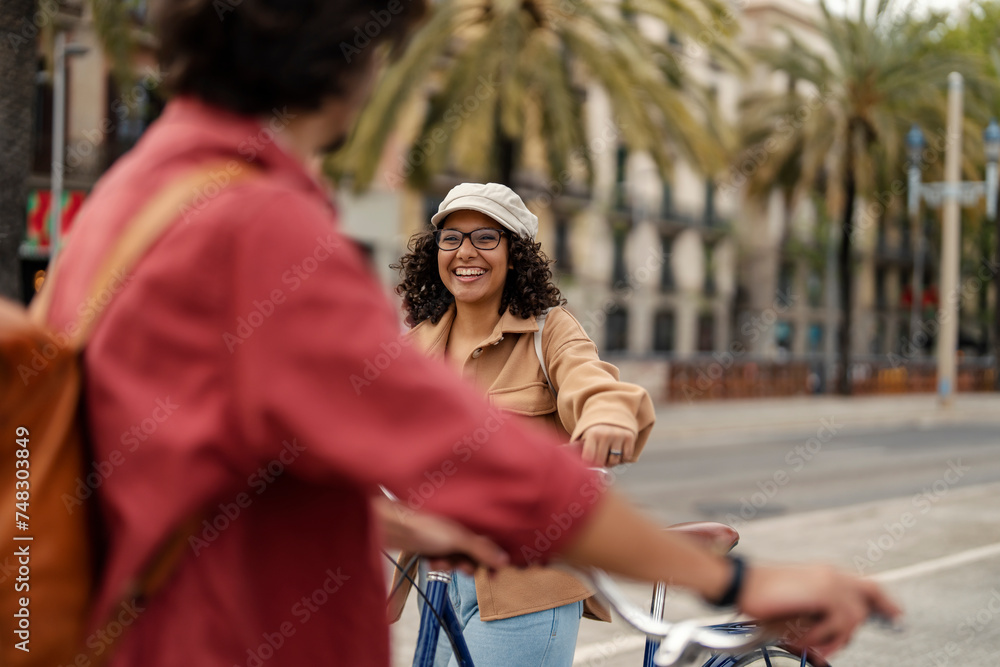 A cheerful trendy girl is pushing bike on the street and meeting a friend.