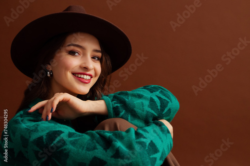 Fashionable happy smiling woman wearing hat, trendy silver hoop earrings, green sweater, posing on brown background. Close up studio fashion portrait. Copy, empty, blank space for text
