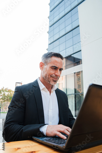 Vertical. Mature business man working with a laptop outside, sitting at coffee shop. Handsome executive male using a computer to operate on the stock market. Successful white collar worker typing