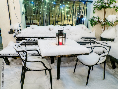 Tables and chairs covered in fresh snow at winter time, burning red candle on the table covered with fresh snow