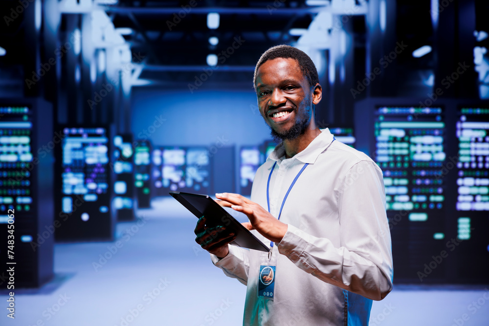 Portrait of african american administrator in front of server clusters in high performance computing data center. Technician monitoring energy consumption across systems, ensuring optimal operations Portrait of african american administrator in front of server clusters in high performance computing data center. Technician monitoring energy consumption across systems, ensuring optimal operations