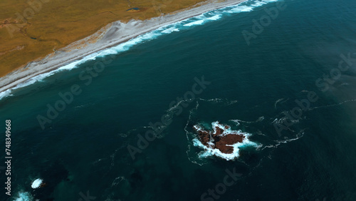 Aerial top view of waves break on rocks in a blue ocean. Clip. Ocean waves crashing against an empty stone rock cliff.