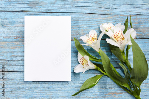 A single white blank card positioned next to white Alstroemeria flowers on a blue painted wooden background