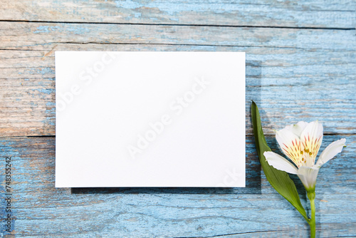 A landscape-oriented blank white card mockup on a weathered blue wooden surface, adorned by a spray of white Alstroemeria flowers