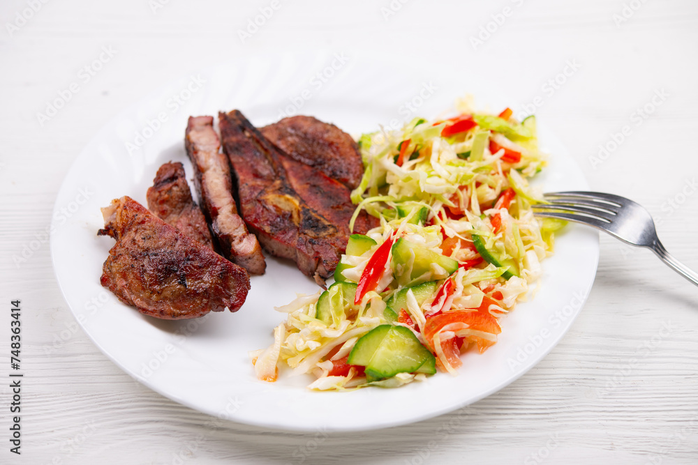 Pork or beef ribeye steak grilled on charcoal. Salad with cabbage, sweet bell peppers, onions, cucumber. Cooked meat and vegetables. Meal. Healthy dinner. Plate on white natural wooden table Pork or beef ribeye steak grilled on charcoal. Salad with cabbage, sweet bell peppers, onions, cucumber. Cooked meat and vegetables. Meal. Healthy dinner. Plate on white natural wooden table