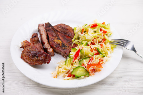 Pork or beef ribeye steak grilled on charcoal. Salad with cabbage, sweet bell peppers, onions, cucumber. Cooked meat and vegetables. Meal. Healthy dinner. Plate on white natural wooden table